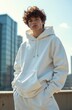 © Viktor - Young man wearing a white hoodie and sweatpants poses with hands in pockets outdoors. Modern city buildings in background. Casual streetwear fashion.