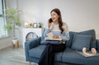 © PHOTO4U - A young woman sits comfortably on a sofa, holding a notebook and smiling while talking on her smartphone. She appears relaxed, multitasking between reading notes and having a conversation.