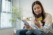 © PHOTO4U - A young woman is sitting on a sofa holding a smartphone and credit card, smiling as she makes an online transaction. She looks relaxed and confident while shopping at home.