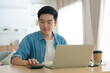 © Monster Ztudio - Happy young Asian man working on laptop in living room. Freelance worker male sitting at desk writing notes while watching webinar, studying online