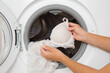 © fotoduets - Young adult woman hand holding and loading white lace bra in mesh sack in opened washing machine. Female delicate under clothes care. Closeup. Front view.