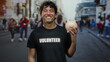© Krakenimages.com - Hispanic man holding piggy bank stands smiling on busy street wearing volunteer shirt showcasing charity and community involvement in outdoor urban environment.
