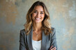 © Vadym - Smiling young woman in grey blazer poses with arms crossed. Female professional with long wavy brown hair and necklace against abstract background. Businesswoman with confident expression.