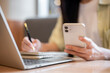 © bongkarn - Close up woman student holding pen writing in notebook looking at phone aside laptop on table in cafe