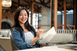 © bongkarn - Smiling asian woman holding pen over notebook and looking away sitting sofa chair at table in a cafe
