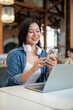 © bongkarn - Happy asian woman with headphones looking at phone while sitting in front of laptop on table in cafe