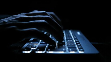 In a dark room a hacker's or programmer's hands are illuminated by the blue light of a backlit laptop keyboard as they type code or breach a system

