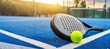 © Chinnawat - A close-up, low-angle shot of a padel racket (or tennis/pickleball racket) and a vibrant yellow ball resting on the bright blue court. The scene is set outdoors during sunset or sunrise, with a warm.
