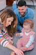 © alfons - Smiling parents sit on the living room carpet watching their curious baby daughter in pink overalls reach for a wooden cup, illustrating playful family bonding,early development and home joy together