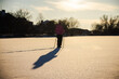 © StockMediaSeller - Woman in a pink jacket walking with trekking poles across a frozen snow-covered lake at sunset. Her long shadow stretches on the glistening snow, symbolizing calmness and winter activity.