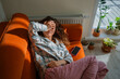 © wifesun - Woman feeling unwell and exhausted while lying on an orange sofa, covering her eyes with a hand, suffering from toxicosis, nausea, or a headache, indicating discomfort and illness