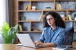 © Liubomir - Young woman with curly hair wearing a headset with a microphone. Concentrating while typing on a laptop. Working remotely from her modern home office setup with a bookshelf and plant in the background