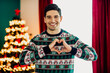 © deagreez - Smiling young man wearing a festive sweater making a hand heart gesture in front of a beautifully decorated Christmas tree