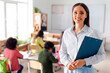 © Home-stock - Portrait of friendly European woman teacher standing in classroom with clipboard during lesson, with pupils sitting at desks on background