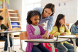 © Home-stock - Friendly European female teacher helping girl pupil during lesson or test in classroom, black schoolgirl writing in notebook, sitting at desk with classmates on background