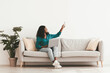 © Prostock-studio - Happy young black woman in stylish casual outfit sitting on couch with laptop at home, holding remote, turning on air conditioner while surfing on Internet, white wall background, copy space