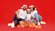© Prostock-studio - Happy Arabic Family Posing With Christmas Gifts Wrapped Boxes Wearing Santa Hats Sitting Over Yellow Background, Smiling To Camera. Winter Holidays And New Year. Studio Shot