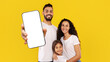 © Prostock-studio - Mobile Offer. Happy Middle Eastern Family Showing Cellphone Blank Screen Recommending Great Application Standing Over Yellow Studio Background, Smiling To Camera. Mockup