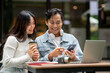 © bongkarn - Young asian couple looking at phone aside laptop sits at outdoor table in front of cafe coffee shop