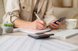 © Nanci - Woman's hands writing notes in a spiral notebook while holding a smartphone, actively managing personal finances, calculating a budget, and planning household expenses on a desk