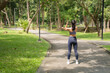 © MINAE - Asian woman standing on a paved path in a lush green park, extending arms across her body, warming up for a fitness workout session and promoting a healthy lifestyle