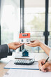 © doidam10 - Close up of real estate agent handing house model to client with calculator and contract paperwork on desk, concept of mortgage loan, home ownership, property investment and housing agreement.