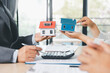 © doidam10 - Close up of real estate agent showing small house model to client with calculator, contract paperwork on desk, concept of mortgage loan, home ownership, property investment agreement. mortgage loan
