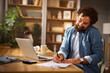 © Stockphotodirectors - A man is seated at a wooden desk in his home office, taking notes while on a call. His laptop is open, with a cozy and well-decorated background creating a productive atmosphere.