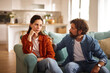 © Stockphotodirectors - Two adults sit on a couch, deep in conversation about their relationship issues. The woman looks concerned while the man offers comfort in a bright and inviting living room.