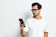 © SHOTPRIME STUDIO - Stylish young man wearing glasses and a casual white t-shirt, gazing thoughtfully at his smartphone, showcasing a modern lifestyle against a minimalistic background.