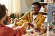 © AnnaStills - Young adult Black man smiling while handing bowl of salad to Caucasian woman during group meal, sitting at dining table with food and drinks, engaging in friendly conversation