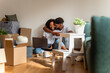 © lordn - African American couple sitting on the floor surrounded by moving boxes, hugging and smiling while using a laptop. Joyful moment of love, teamwork, and starting a new home together