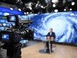 © Moopingz - Professional broadcast camera on a tripod in a TV studio focusing on a weather report with a hurricane map.