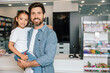 © zinkevych - Bearded father in casual denim shirt holding daughter smiling together in modern pharmacy
