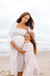 © maxbelchenko - A young pregnant woman hugs her daughter on a sandy beach. A beautiful woman and her child walk along a sandy beach, enjoying time together. The concept is about family, childhood, and fun.