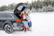 © maxbelchenko - Two joyful women in cozy clothes sit on a blanket in the trunk of a car on a winter day. Beautiful friends enjoy a car trip through a snowy forest. Concepts of friendship, fun, and seasonality.