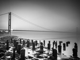 Black and white image of the Humber Bridge spanning calm water at sunrise with wooden posts in the foreground