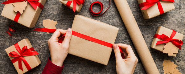Wall Mural - Woman is packing surprise gift for Christmas holidays, sitting at workplace with craft tools, top view. Woman wrapping Christmas gift at colored table