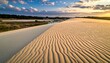 © Sapuy - A scenic landscape portrays a sand dune with textured ridges under a cloudy sky at sunset. Vegetation borders a body of water in the distance