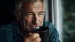© Bussakon - A thoughtful mature man holds a coffee mug and gazes out a window in soft natural light