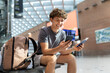 © Anastasiya - Smiling traveler guy sitting at airport bench using smartphone and holding passport. Happy young man waiting for boarding or booking hotel abroad