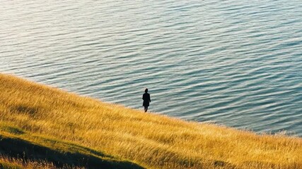 Wall Mural - A person stands at the top of a hill overlooking a serene body of water, perfect for use in travel, nature, or outdoor-themed projects