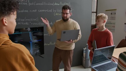 Medium zoom-out shot of enthusiastic young Caucasian male coding teacher, holding laptop, demonstrating computer script to multiethnic students on screen during web design lesson at school