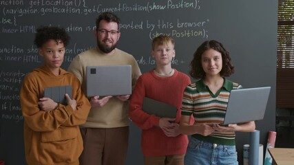 Medium portrait of happy diverse young male coding teacher and teenage students, holding and gadgets, posing together in front of blackboard with computer script at web design lesson in middle school