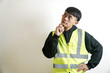 © yashis007 - Portrait of a thoughtful young Asian construction worker in a reflective safety vest, contemplating a problem or planning a strategy against a neutral background