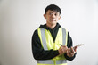 © yashis007 - Asian man wearing a high visibility vest writing on a clipboard, looking at camera with a neutral expression, isolated on a white background