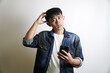 © yashis007 - A confused young Asian man in a denim jacket holds a smartphone and scratches his head while looking at the camera, questioning a concept or idea
