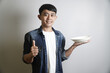 © yashis007 - Young Asian man holding a white plate and giving a thumbs up gesture with a smile