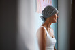 © DragonImages - Middle aged Caucasian woman wearing headscarf sitting in medical clinic, facing sideways, showing signs of cancer treatment, medical poster visible on wall in background