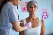 © DragonImages - Middle aged Caucasian woman wearing headscarf sitting while female healthcare worker examining chest with stethoscope, cancer awareness posters visible in background